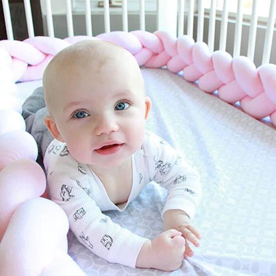 Jeune bébé souriant dans son lit à barreaux avec un tour de lit tressé rose gris et blanc