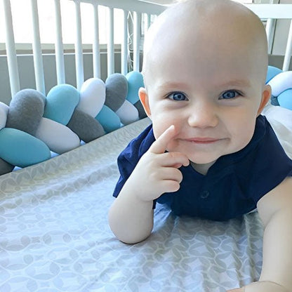 Jeune bébé souriant dans son lit à barreaux avec un tour de lit tressé bleu gris et blanc